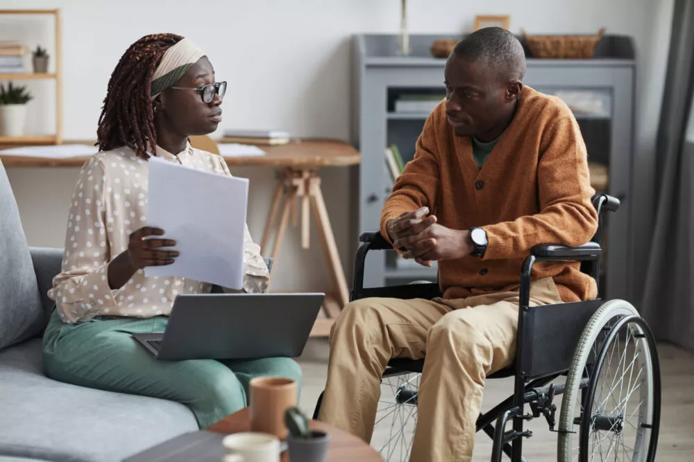 un homme en fauteuil roulant discute dans son salon avec une femme qui lui montre des documents le concernant