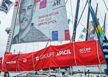 Le bateau de Damien Seguin en mer, coque rouge, la voile rouge et blanche avec le portrait du navigateur, mention Apicil sponsoring